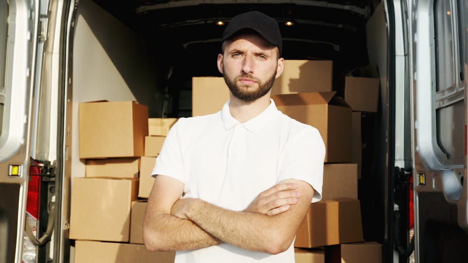 A man wearing a face mask, grey trousers, a beige and brown vest, and a cap is standing beside a white delivery van on a paved driveway, positioned next to a brick pillar and a metal gate. The van is