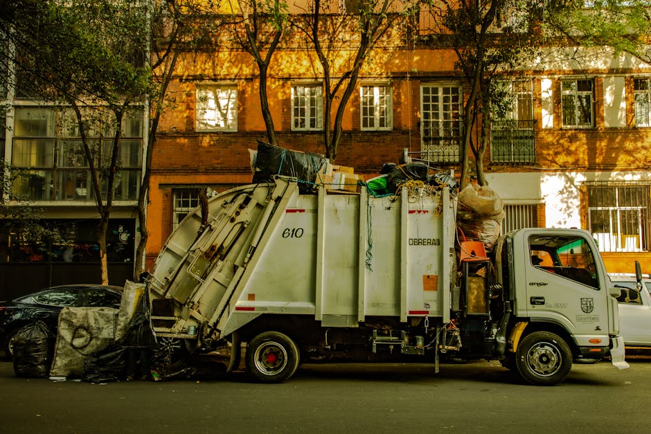 A white garbage and recycling collection vehicle with the number 610 on its side, parked on a residential street during daylight, is shown in the process of collecting household waste and bulky items for disposal. The vehicle, branded with 'O'DREIRAL,' is loaded with various discarded objects, including large plastic bags, cardboard boxes, and bulky furniture such as a dismantled or broken piece of wooden furniture covered in protective material. The loading process takes place near a red-brick building with multiple windows, some with white frames, and the premises are shaded by tall trees along the sidewalk. Nearby, additional cars are parked along the street, and the scene is illuminated by natural sunlight. The image reflects the logistics involved in home relocation or furniture transport, with materials and equipment indicating waste collection or clearance activities, which are typically handled by companies like Man with Van Morden as part of their removals and disposal services.