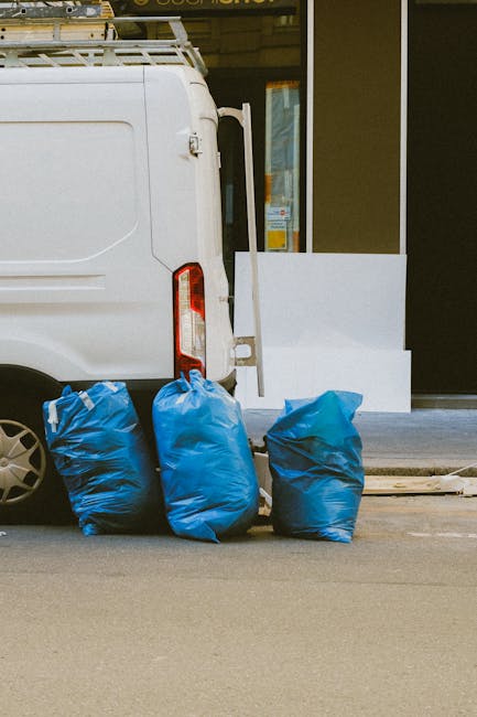 A portion of a white delivery van with its side door slightly open, positioned on a street in front of a building with a glass window. Three large blue plastic bags filled with bulky waste or sofa disposal materials are placed on the pavement next to the van, ready for loading or unloading. Inside the van, visible packing materials include cardboard and possibly fabric covers, indicating ongoing home relocation or furniture transport. The scene suggests a professional removals process involving loading or disposing of large items, with the van aligned for efficient transportation. Man with Van Morden, a company specializing in removals, appears to be engaged in a packing and moving activity, supporting services like bulky waste disposal during house removals or furniture transit. The environment is well-lit, typical of a daytime operation, with an emphasis on proper handling of large household items.