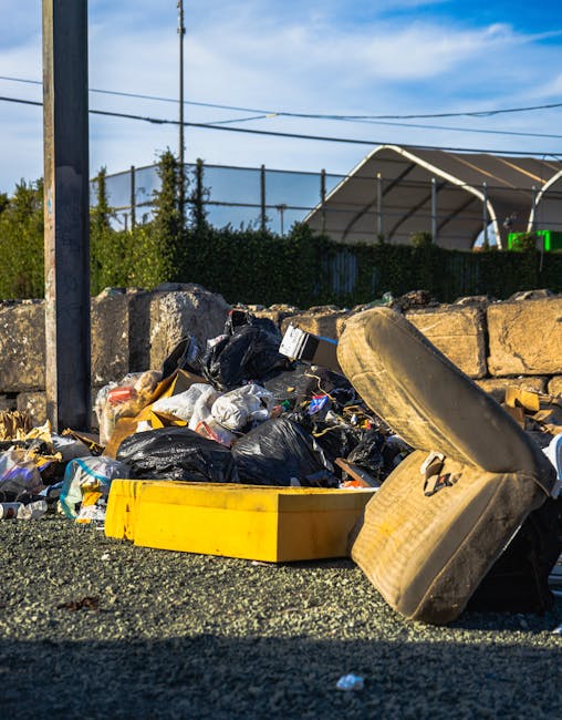 A white garbage and recycling collection vehicle with the number 610 on its side, parked on a residential street during daylight, is shown in the process of collecting household waste and bulky items for disposal. The vehicle, branded with 'O'DREIRAL,' is loaded with various discarded objects, including large plastic bags, cardboard boxes, and bulky furniture such as a dismantled or broken piece of wooden furniture covered in protective material. The loading process takes place near a red-brick building with multiple windows, some with white frames, and the premises are shaded by tall trees along the sidewalk. Nearby, additional cars are parked along the street, and the scene is illuminated by natural sunlight. The image reflects the logistics involved in home relocation or furniture transport, with materials and equipment indicating waste collection or clearance activities, which are typically handled by companies like Man with Van Morden as part of their removals and disposal services.