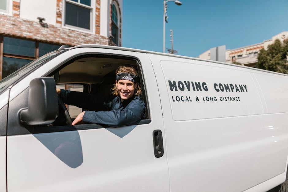 A smiling male mover with long hair and wearing a headband is sitting inside the cab of a white moving van parked outside a residential building. The van displays the text 'MOVING COMPANY' along with 'LOCAL & LONG DISTANCE' on its side. The driver is looking out of the open window, with his arm resting on the door, demonstrating a friendly and professional approach to home relocation and furniture transport. The background includes a building with large windows, a clear blue sky, and some greenery. The scene illustrates the loading process, possibly preparing for packing and moving services, using equipment like blankets or straps might not be visible but is implied for safe furniture transport by [COMPANY_NAME], which offers quick and reliable removals services in the Morden area.
