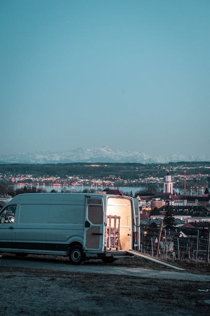 A silver panel van with its side door open, parked on an elevated area overlooking an illuminated cityscape at dusk. Inside the van, a wooden furniture piece with visible legs and a flat top is being loaded or unloaded, supported by a ramp extending from the vehicle to the ground. Adjacent to the van, on the ground surface made of gravel or asphalt, are cardboard boxes and packing materials, indicating a home relocation or furniture transport process. The background features a sprawling urban area with numerous buildings, some with lit windows, and a distant mountain range faintly visible under a clear sky. This scene reflects a professional moving service focusing on last-minute or same-day house removals, consistent with the service offerings of Man with Van Morden.