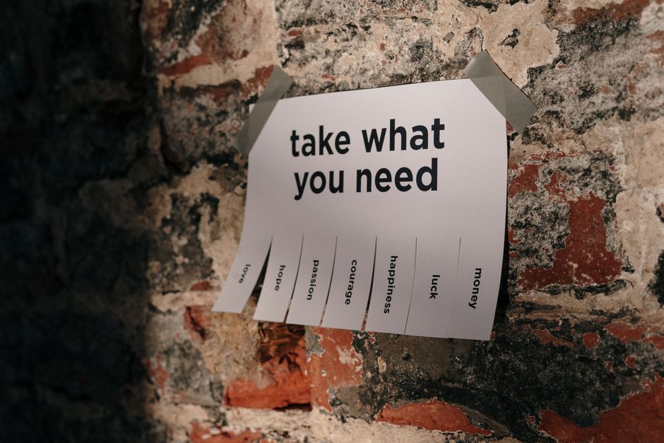 A printed sheet titled 'take what you need' is taped to a rough, weathered brick wall with grey duct tape at each corner. The sheet displays several vertical tabs, each with a different word including 'hope,' 'passion,' 'courage,' 'happiness,' 'luck,' and 'money,' with one tab labeled 'hope' partially torn away. The background wall has a mix of dark and reddish bricks with visible mortar, indicating a rustic or industrial environment. The image captures an indoor setting that could be part of a home or storage area, with the sheet possibly serving as a motivational or organizational aid related to home relocation or packing tasks. The presence of this motivational note in a space associated with moving reflects the themes of packing and mental preparation involved in house removals, as handled by companies like Man with Van Morden.