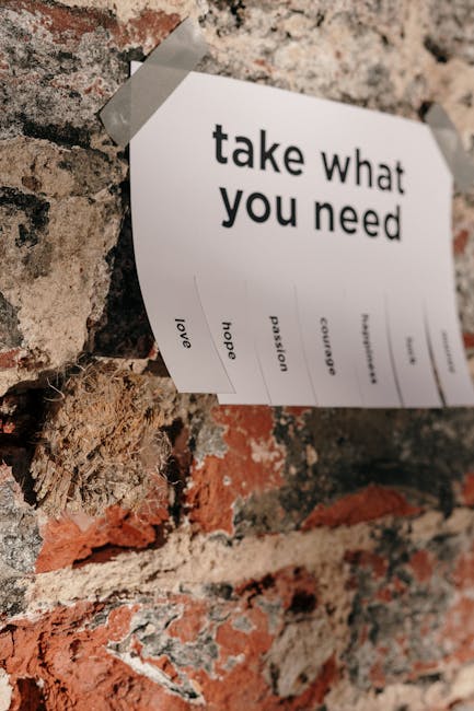 A close-up photograph of a printed sheet of paper taped to a textured, weathered brick wall, with the phrase 'take what you need' prominently displayed in bold black letters at the top. Below the main message, there are smaller words such as 'love', 'hope', 'passion', and 'courage' arranged vertically, suggesting offerings of motivational or supportive sentiments. The paper appears to be attached with clear tape at the top corners, and the wall's surface shows a mix of reddish-brown and gray bricks with rough, uneven mortar. The setting evokes a sense of casual, accessible communication, possibly in an urban or residential environment where messages of kindness are shared openly. This scene aligns with themes of community, support, and movement, relevant to home relocation and moving services, subtly reflecting the ethos of careful handling and positive encouraging spaces, as seen with [COMPANY_NAME] in the context of house removals in Morden.