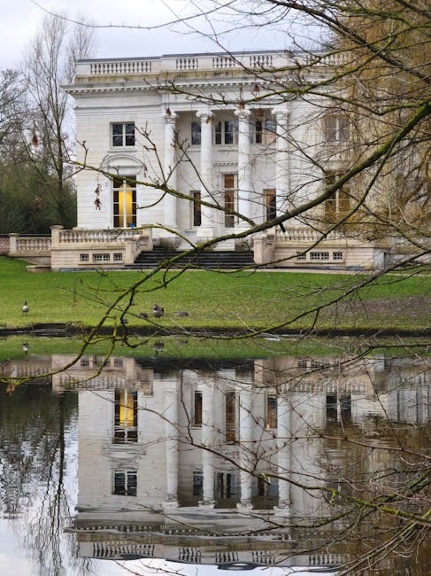 A white, neoclassical-style house with tall columns and decorative balustrades on the roof sits behind a grassy area and a body of water that reflects its facade. The scene is framed by leafless tree branches in the foreground, indicating a winter or early spring setting. The house's windows are illuminated, suggesting interior lighting. The presence of a stone staircase leading to the entrance and the lush green lawn in front imply this is a residential property. In context with the house removals and relocation services offered by Man with Van Morden, staff may be preparing to load furniture and boxes into a nearby vehicle, visible on the pavement or driveway, for safe transport. The environment appears calm and organized, suitable for a home relocation process involving careful packing and furniture transport, supported by professional removalists.
