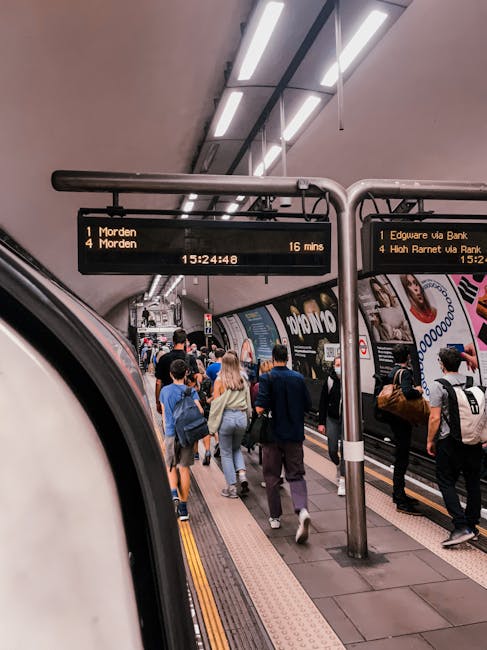 A crowded underground train station platform with passengers walking towards waiting trains. The platform features an electronic timetable display overhead, showing departure times and destinations, i