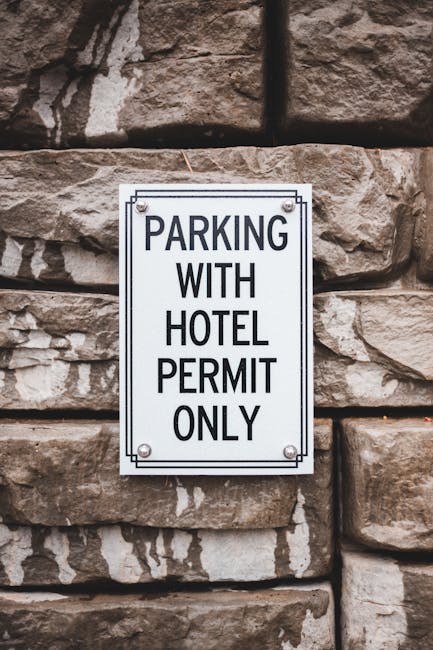 A rectangular metal sign mounted on a rough stone wall, displaying the text 'PARKING WITH HOTEL PERMIT ONLY' in bold black capital letters. The sign is secured with four screws, one at each corner. The wall behind the sign consists of large, uneven sandstone blocks with visible mortar joints. The scene appears to be outdoors, possibly near a building entrance or parking area, with natural light illuminating the textured stones. This signage may be relevant to vehicle restrictions during a home relocation or furniture transport process managed by Man with Van Morden, especially when parking permits are required for efficient loading and unloading outside the property.