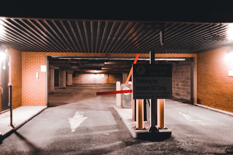 The image shows the entrance to an underground parking garage with a brick wall on both sides and a dark, ribbed ceiling overhead. A sign mounted on a black pole indicates parking restrictions, mentioning parking lot 60 and permit requirements, with the text partially obscured. A red and white barrier arm is lowered across the entrance, blocking vehicle access. In front of the barrier, there are two concrete bollards and a small concrete island. The pavement in front of the entrance is marked with a white arrow pointing towards the garage, indicating the direction of traffic flow. To the left, part of a black gate or door is visible, and the overall environment suggests a secure, commercial or residential parking area, suitable for home relocation or furniture transport logistics handled by Man with Van Morden. The lighting inside the garage is dim, with artificial lights illuminating the area, emphasizing the barriers and signage used for managing parking and access control during moving operations.