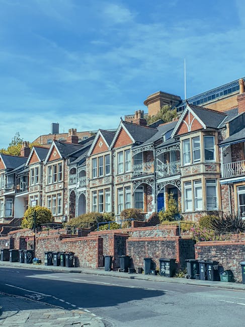 A row of traditional Victorian terraced houses with ornate front facades, decorative ironwork balconies, and bay windows, situated on a slightly elevated street with brick retaining walls and landscaped front gardens. The houses are constructed of brick with painted embellishments, and the area features a clear blue sky with scattered clouds. In the foreground, black wheelie bins are lined along the pavement, indicating residential waste collection. The scene captures a quiet residential neighbourhood during daytime, highlighting classic UK home architecture relevant to house removals and relocation services provided by Man with Van Morden, supporting home relocation and furniture transport processes.