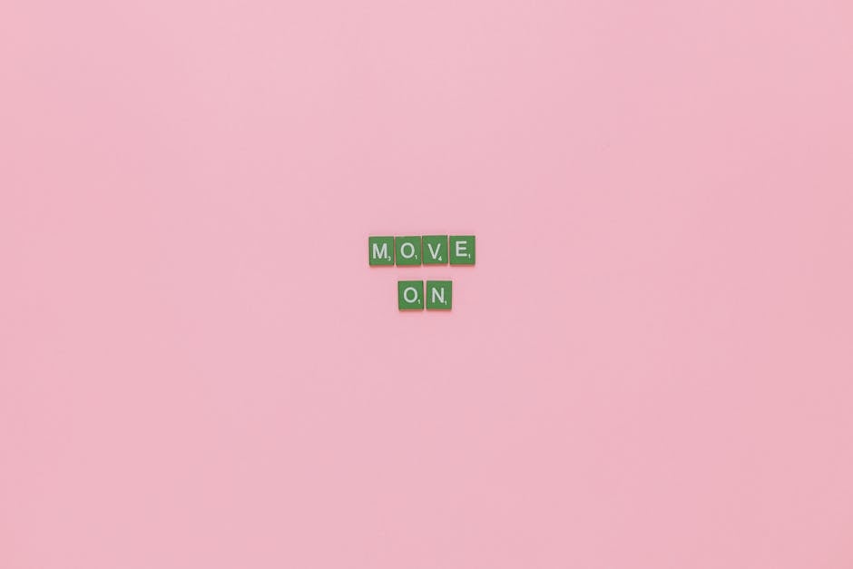 An image showing small green square letter tiles arranged on a pastel pink background spelling out 'MOVE, ON'. The tiles are placed in a scattered, casual manner, with the word 'MOVE,' positioned above 'ON'. The background is plain, providing no additional objects or environment details. This visual could relate to themes of moving or transition, which supports the context of home relocation and furniture transport services offered by companies like Man with Van Morden, especially on a page about transparent removal quotes in Morden. The overall composition emphasizes the message of moving forward or starting anew.
